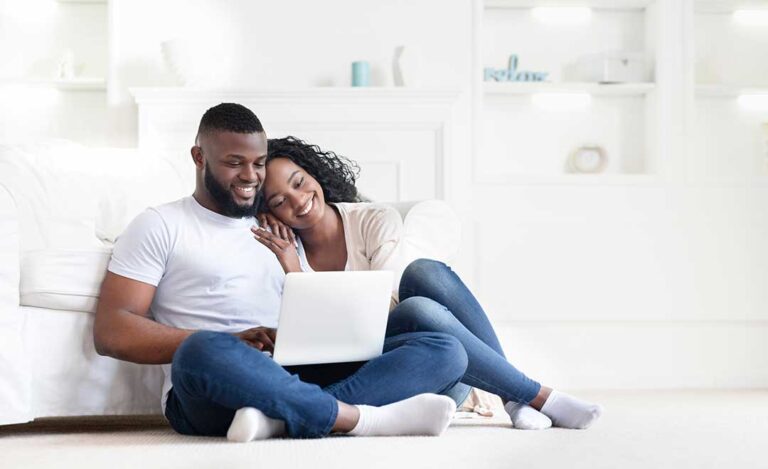 A couple reviews paperwork at a kitchen table with a laptop and home documents spread out.